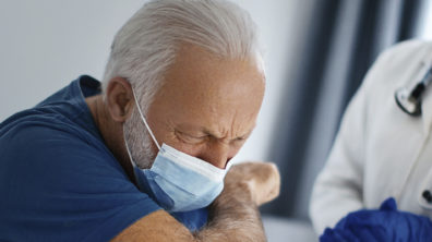 Senior patient sitting on the bed, coughing under protective face mask.
