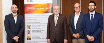 Der Moderator und drei Redner stehen neben einem Roll-up, das das Symposium ankündigt